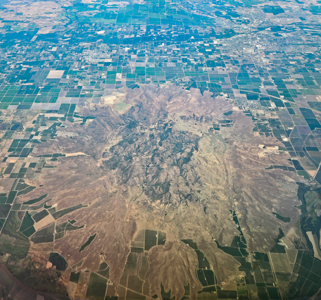 A photo taken from an airplane at high altitude shows a cluster of craggy, brown and gray volcanic peaks surrounded by a circular apron of sloping, vegetated debris. Surrounded the volcano are green and brown agricultural fields laid out in a grid pattern.