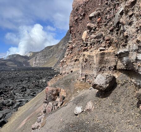 Color photograph of explosive volcanic deposits in wall of crater with degassing volcanic vents in the background