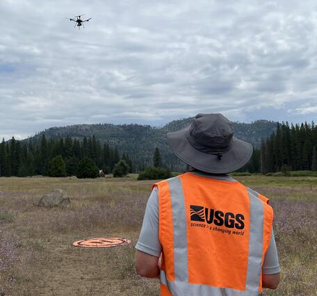 UAS pilot wearing USGS safety vest operates craft hovering above landing pad located in mountain clearing