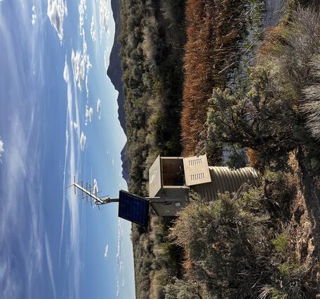Tan solar-powered monitoring station in shrubland near ponding water with mountains under partly cloudy blue sky.