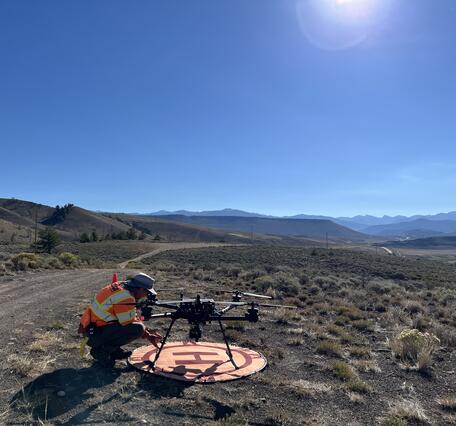 USGS drone pilot squatting by craft on landing pad located along dirt road with mountains in the distance.