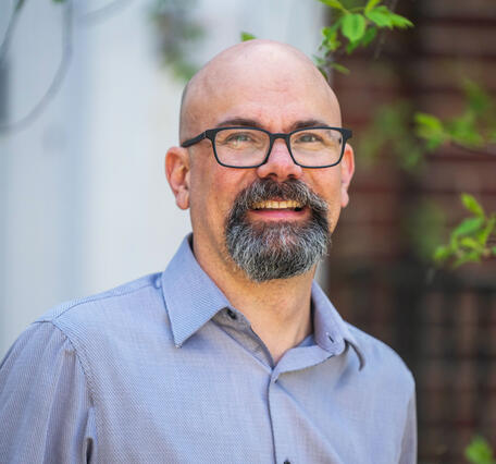 Man wearing a blue button-down shirt standing outside with tree behind him