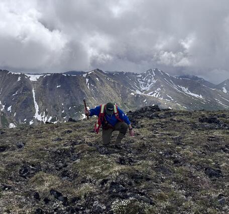 A man kneeling and holding a hammer on an alpine ridge.