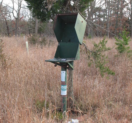 A well standpipe with an open access box on top stands in the middle of a grassy field
