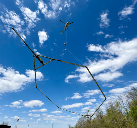 Looking up at a helicopter in flight with scientific instrument under it against a blue sky with white clouds