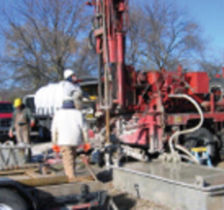 Several people are seen standing near a large underground drilling rig. 