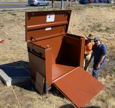 two men in hat and glasses stand by a large brown metal box that is taller than them. In a ditch by a roadway.