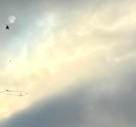 The silhouette of a helicopter with scientific instrument against a grey, cloudy sky 