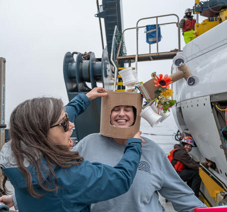 Two people on the deck of a boat laugh while one person wears a man-made hat