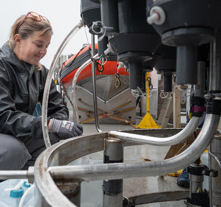 A person smiles while sitting on the deck of a boat as they collect water from tubes coming out of a oceanographic instrument