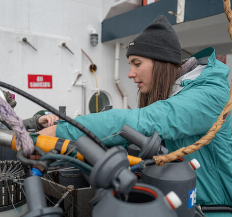 A person in a hat and teal jacket prepares Niskin bottles on a CTD