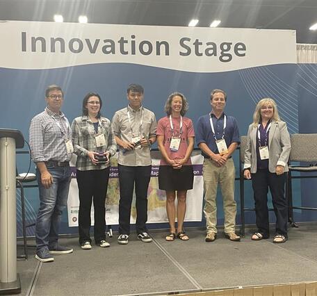 Photograph of six people on a stage with a blue background and high banner reading "Innovation Stage."