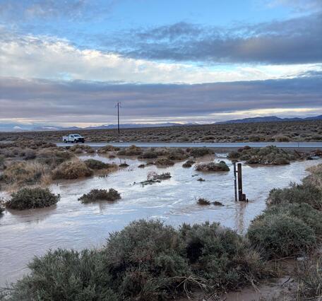 A white USGS truck is driving on a road behind a swollen river surrounded by sagebrush. It is dusk with the mountains silhouetted in the background.