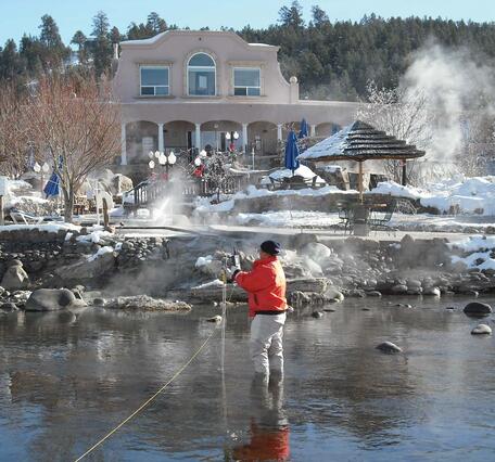 Female wearing orange float coat and black hat stands in calf deep water. Steam from hot springs visible.