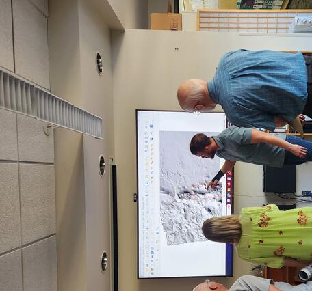 A man in a green shirt points to a planetary geologic map on a screen while engaging with the public at Astrogeology