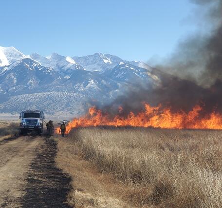 People standing near a fire, white truck, and snow-capped mountains in the background 