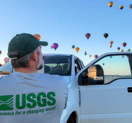 A USGS employee standing in front of their work vehicle looks at the dozens of hot air balloons rising in the sky