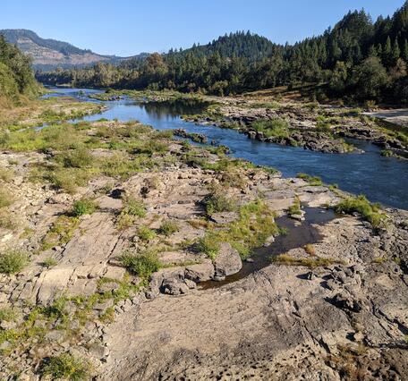 a wide, rocky riverbed with shallow, clear water in the foreground. Rolling forested hills and blue sky in background