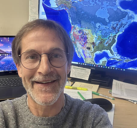 Selfie portrait of Cal Ruleman in front of computer showing geologic map of North America