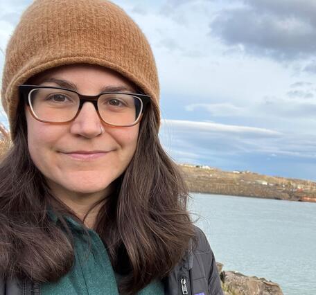 Dark haired female in a tan beanie and glasses stands near the Columbia River on a cloudy day