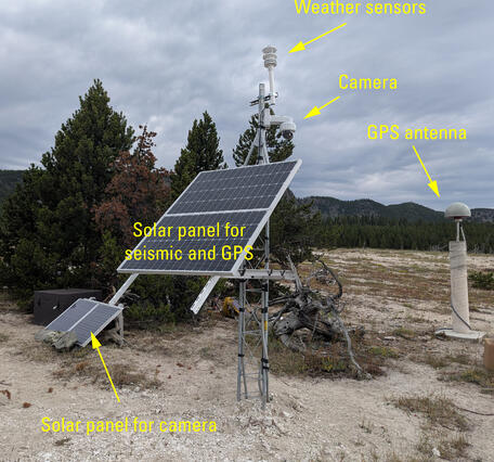 barren ground with a few small pine trees and a metal tower with a solar panel and other sensors