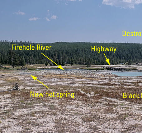 remote camera view of barren ground with a blue hot spring in the middle distance and a forested ridge in the background