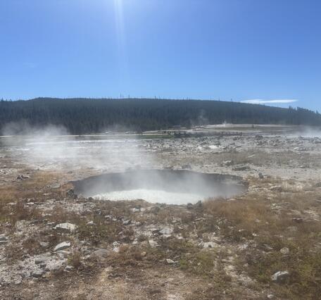 Circular pool of boiling water in a barren area strewn with rocks with a forested ridge in the distance under blue sky.