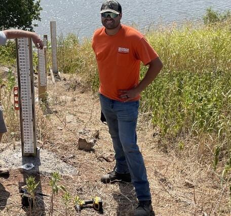 Man with orange USGS shirt, jeans and baseball hat standing at water front