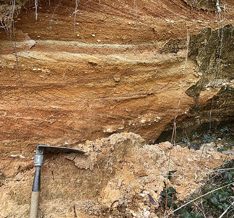 An orange and yellow outcrop of Cretaceous sand and gravel with a digging tool propped up against a bank