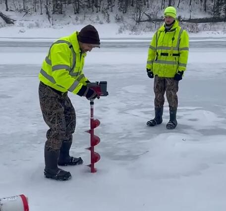 Two scientists standing on a frozen river drilling a hole to check the flow below