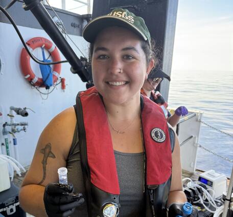 Woman in a grey shirt and red life jacket smiling at the camera while holding a few scientific collection tubes.