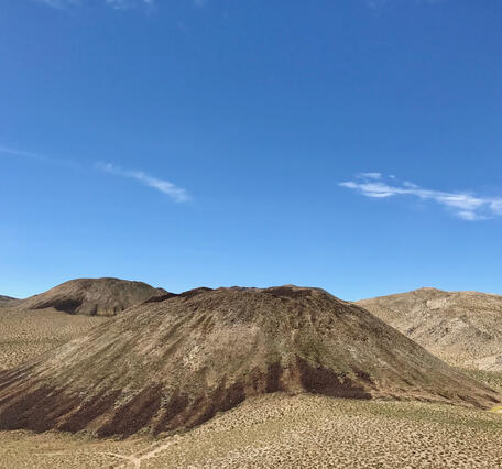 A photograph of several rocky volcanic hills shaped like upside-down flans. The hills and the landscape around them are dotted with sagebrush and dry grasses, revealing the underlying desert soil and rock. A bright blue sky overhead is streaked with clouds. USGS photo by Seth Burgess