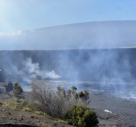 Hazy view of a crater wall with some vegetation in the foreground and a shield-shaped mountain in the background