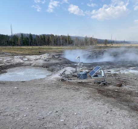 Barren ground with some equipment and solar panels in front of steaming pools.  Grassy meadow and forested area behind.