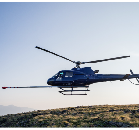 Blue helicopter with boom protruding in front, silhouetted in flight against the sky, rotors frozen in time.