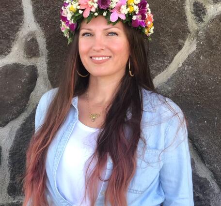 Person with long dark hair with a blue shirt in front of a stone wall wearing a flower crown lei