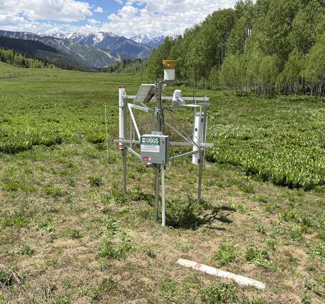 CRNS station in grassy meadow with trees and snow‑capped mountains under cloudy sky.