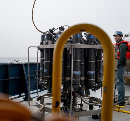 Two people in hard hats and life jackets stand on the deck of a research vessel next to the CTD