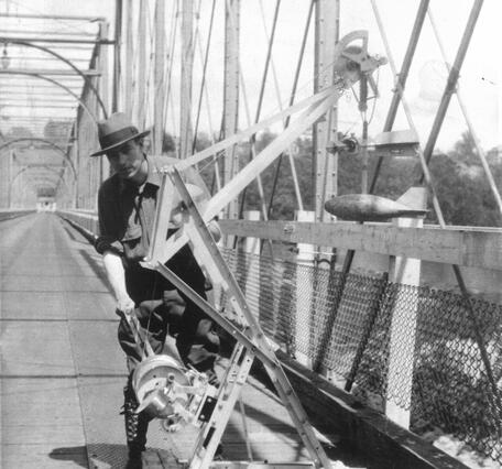 A black and white photo at the Chain Bridge along the Potomac River showing a technician posing next to a boom crane.