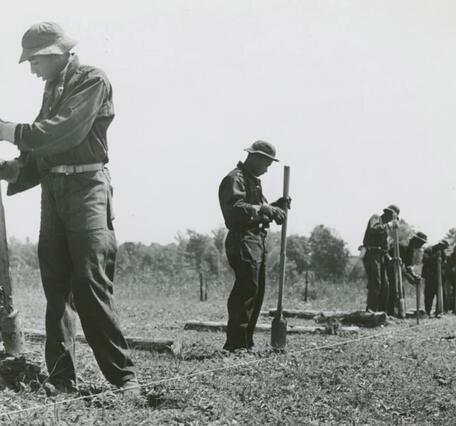 A historical image showing the Civilian Conservation Corps labor force wearing dark work overalls holding hole diggers in preparation of fence pole placement.