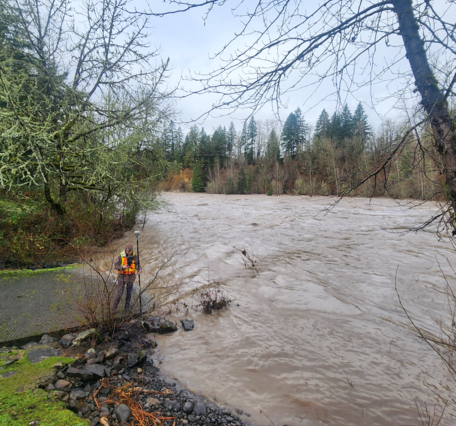 brown Clackamas River flood waters reach upper boat ramp. Drowned trees line the full river in the distance.