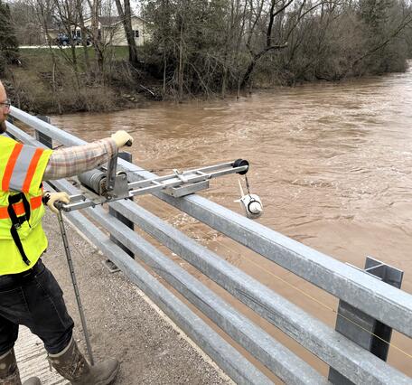 Hydrologic technician collects water sample from flooded river while standing on a bridge