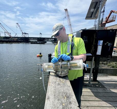 A man in a cap, neon yellow shirt, and blue gloves holds a Van Dorn sampler on a dock with a streamgage and bridge behind.