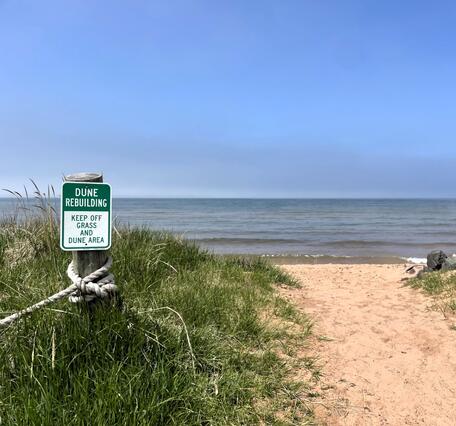A sandy path leading to water, with a sign on the left that states "Dune Rebuilding, Keep Off Dune and Grass Area"