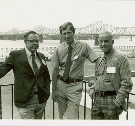 A sepia toned image shows scientists from the National Fishery Research Laboratory in 1981 standing along a metal railing with a large bridge in the background