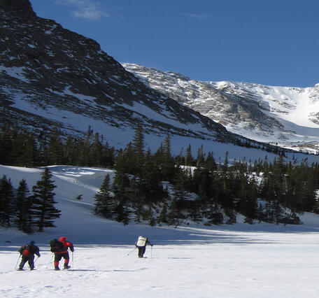 Image of two scientist trekking through snow with Rocky Mountains and trees in background