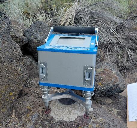 Box-shaped electronic instrument with a digital screen set up on rocky terrain surrounded by dry vegetation and mountains in the background.