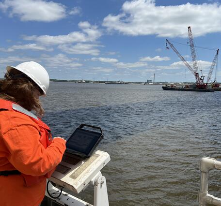 Scientist with a computer set on the bridge rail, looking out over the Delaware River at a crane barge in the distance 