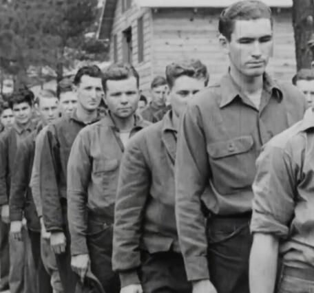 a historical image showing a line of young men in the Civilian Conservation Corps with a large wooden cabin and woods in the background