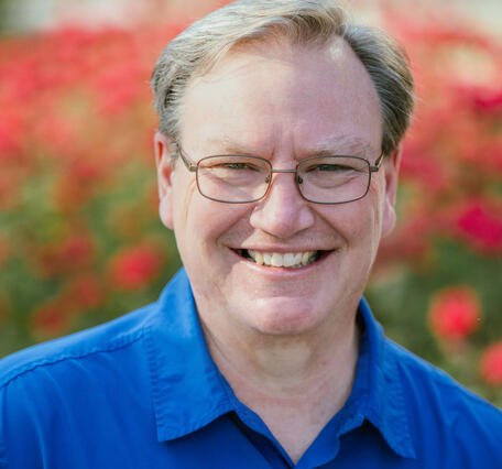 A man in a blue collared shirt wearing glasses and seated in front of red flowers.
